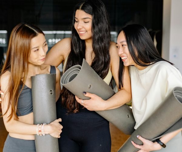 Three women smiling and holding yoga mats during a fitness class indoors, embracing friendship and wellness.